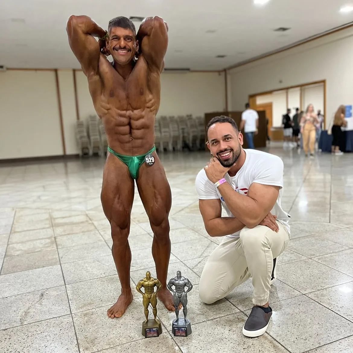 Franthesco Curbani agachado ao lado de um atleta de fisiculturismo e seus troféus